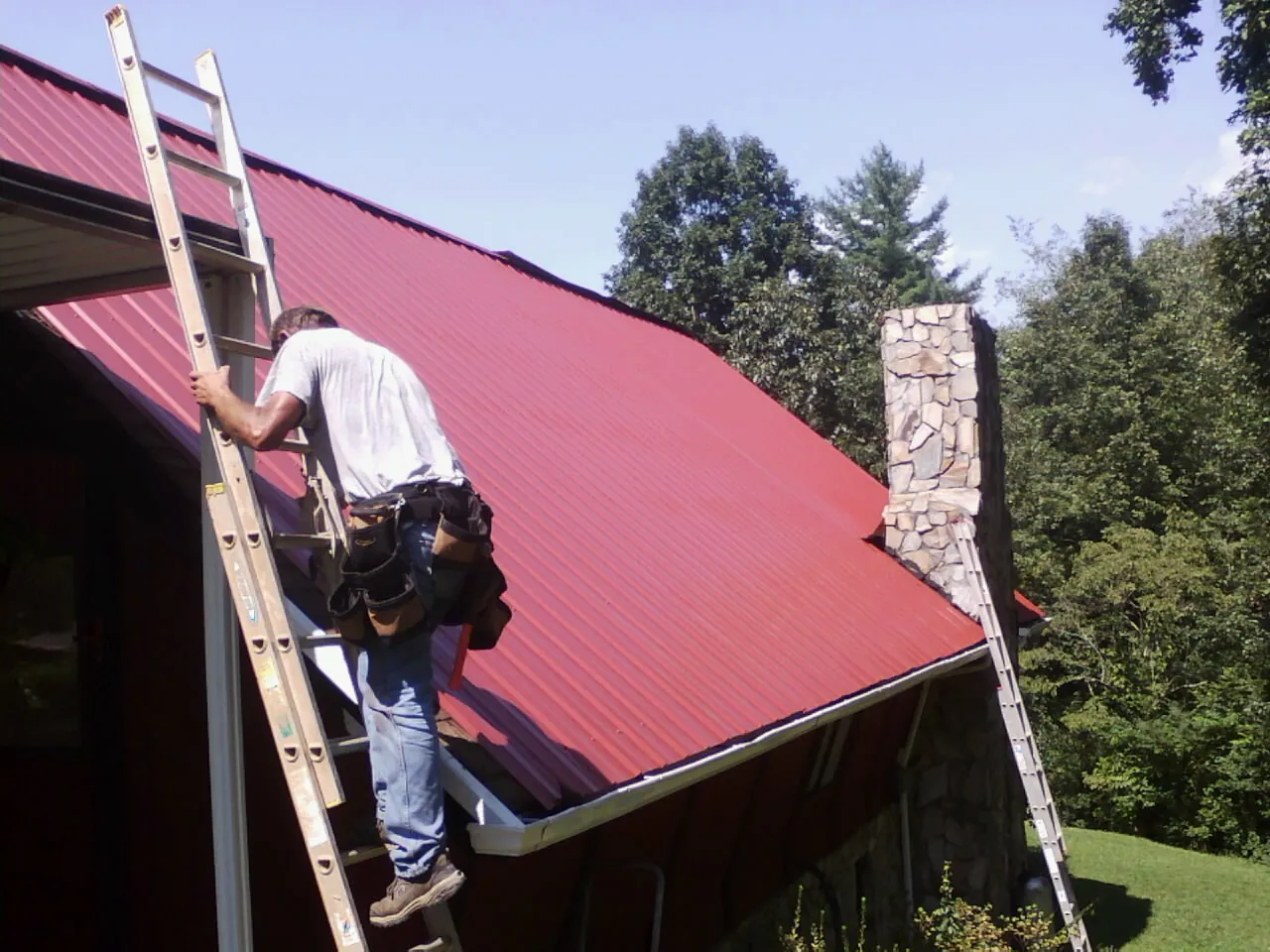 Roofer on a ladder beside a red metal roof and stone chimney — Soffit & Fascia Repair by Roofing Brothers in Paton